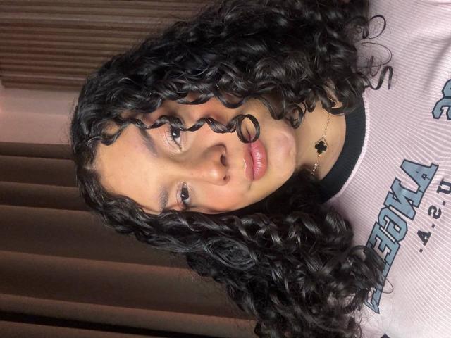 RachellBeaumont has curly black hair, light brown skin, and is wearing a pink shirt with a black necklace. She's looking at the camera with a slight smile.