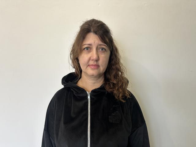RoseHille, middle-aged woman with wavy brown hair, wearing a black zip-up hoodie, standing against a plain white wall, has a neutral expression and slightly pursed lips.