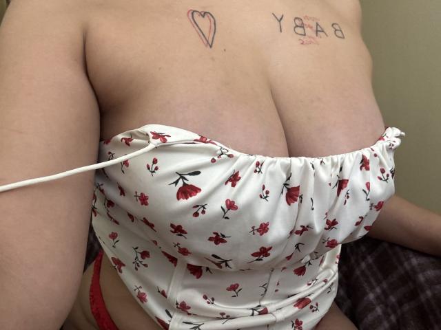 Kametia's big tits are exposed, wearing a white floral top pulled down, showing red lace panties, with tattoos on her chest. Her skin is pale, and she's sitting.