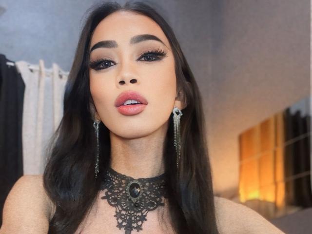 AnastahaDimitri has long, straight black hair and a light olive skin tone. She wears dramatic, dark eyeliner and long, thick, black eyelashes. Her lips are slightly parted with pink lipstick. She has a black lace choker with an ornate, oval brooch in the center. Her earrings are long and silver with a skull design. The background is blurry, with white curtains on the left and a warm light source on the right. The image is a close-up portrait.