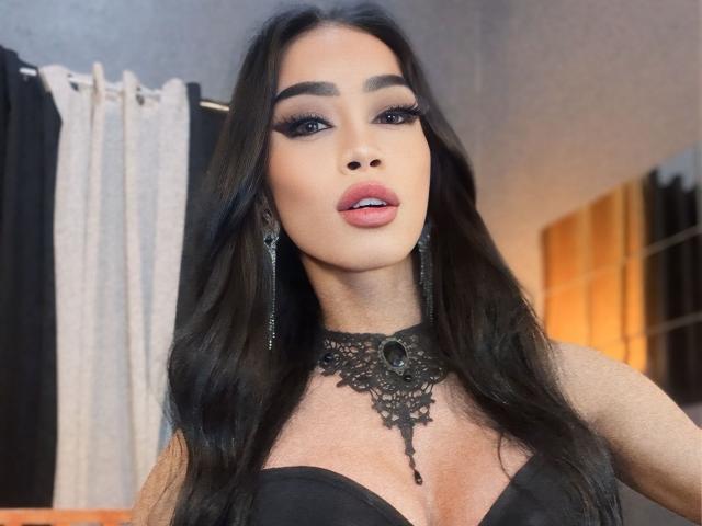 AnastahaDimitri with long black hair, dark eyes, and pink lips, wearing a black lace choker and dress, showing her cleavage and looking at the camera.