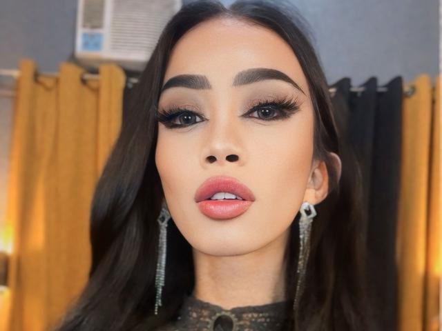 Close-up of AnastahaDimitri with dark, straight hair falling over her shoulders. She has thick, arched eyebrows, long, heavy black eyelashes, and subtle eyeshadow. Her lips are full with a pinkish-red hue. She wears silver, teardrop-shaped earrings and a black lace top. The background shows yellow curtains and a black curtain to the right. The image is sharply focused on her face, with warm, soft lighting. Her expression is neutral, with slightly parted lips. The texture of her hair is smooth and shiny.