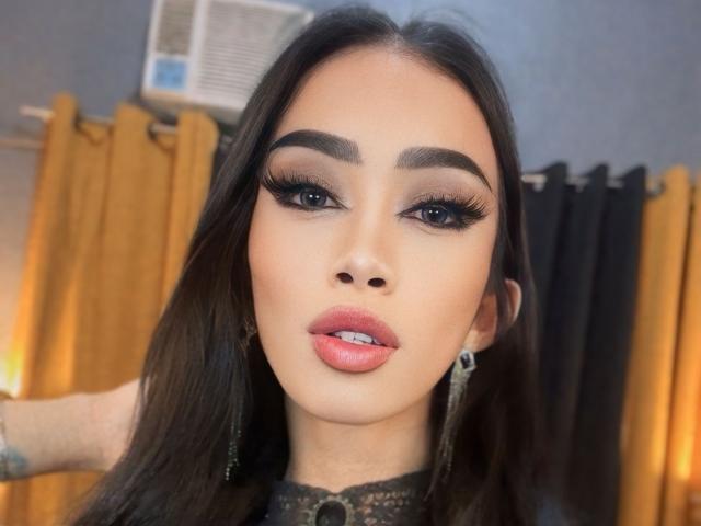 AnastahaDimitri has dark brown hair, thick eyebrows, long eyelashes, and pink lipstick. She's wearing black lace earrings with a yellow curtain in the background.