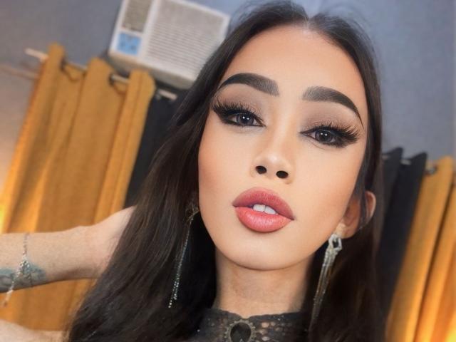 AnastahaDimitri with dark hair, heavy eyeliner, full pink lips, and a tattoo on her wrist, wearing long earrings, in a yellow curtain room. Air conditioner above.