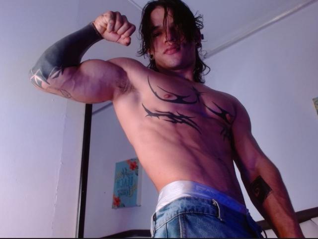 Der muskulöse AlexBernal mit schwarzen Tattoos auf Brust und Arm, der seinen Bizeps anspannt, trägt blaue Jeans, weiße Unterwäsche und einen schwarzen Armärmel, hat langes, dunkles Haar und ein leichtes Lächeln..