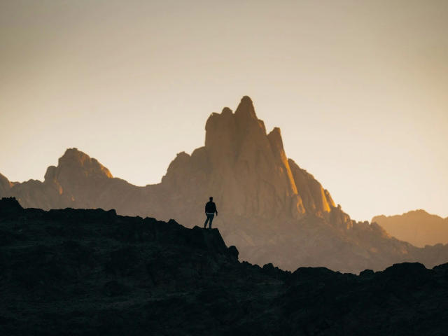 Silhouetted rs03o7Test12-hot stands on a rocky outcrop, facing a jagged mountain peak bathed in sunset light. The mountain's sharp, uneven edges contrast with the smooth gradient of the sky, which transitions from warm yellow to cool gray. The dark rocky foreground emphasizes the mountain's illuminated, textured surface. The composition uses strong, natural lines and creates a dramatic, elevated perspective. The overall tone is warm and majestic.