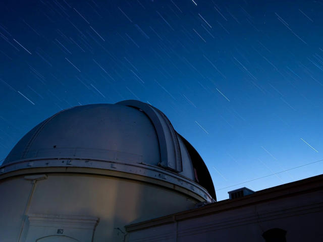 A night sky photograph with long-exposure star trails. A large, dome-shaped telescope takes up the lower half, its metallic surface reflecting minimal light. The dome's texture is smooth, with subtle, darker lines indicating wear. The star trails, diagonal and bright, dominate the upper half, creating a dynamic contrast against the dark blue sky. The telescope's lower part is illuminated, revealing a small, rectangular light source near the bottom right. The composition emphasizes the telescope against the moving star trails.