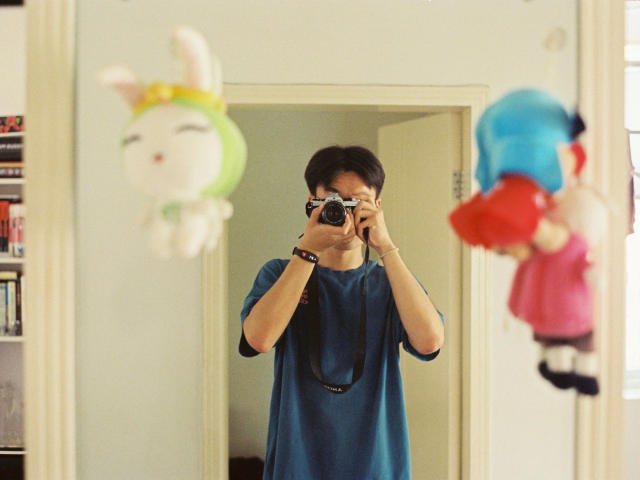 rs0307Test1234-hot in a blue shirt, holding a camera, taking a mirror selfie, with two plush toys, one with green and white, and one with blue and red, hanging.