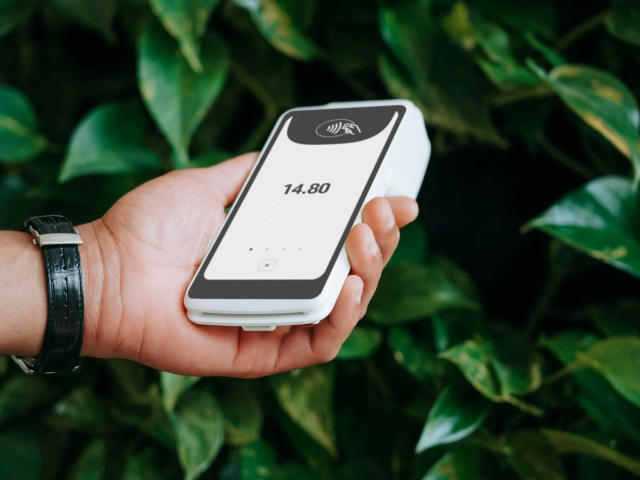 A close-up of a hand holding a white, rectangular smartwatch with a black screen. The screen shows "14:30" in black text. The hand has a light skin tone and wears a black leather wristwatch with a silver buckle. The background is lush green foliage. The smartwatch is centered, and the hand is slightly angled to the right. The image focuses on the smartwatch and the hand.