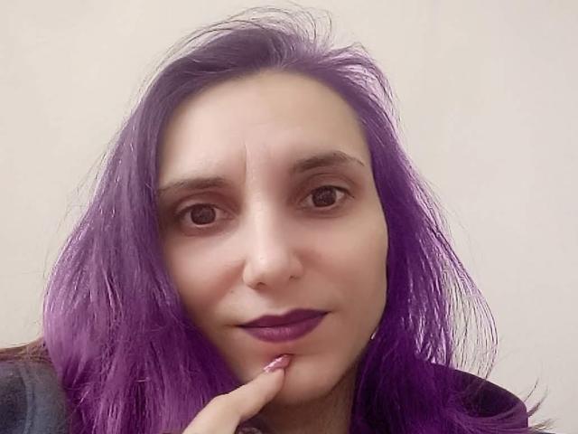 Close-up of PussyWetMistress with purple hair, light skin, and dark purple lipstick. She's looking at the camera with a slight smile, her right index finger touching her lower lip. Her hair is messy and falls around her face. She has brown eyes and light eyebrows. Her expression is neutral but slightly curious. The background is a plain, light beige color. Her nails are painted purple, matching her lipstick. The image has a slightly overexposed, soft look. The focus is on her face and hair.