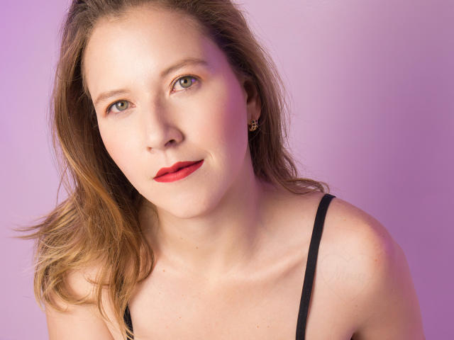 LyraBloom has fair skin, red lipstick, and brown hair, wearing a black strap top, with green eyes and small gold earrings, looking at the camera with a slight smile.