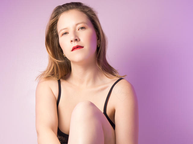 LyraBloom hat helle Haut, roten Lippenstift und hellbraunes Haar, trägt schwarze Dessous und hat ein Knie angezogen, wobei sie leicht zur Seite schaut, vor einem rosa-lila Hintergrund..