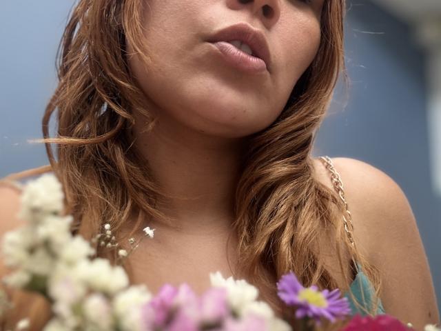 Close-up of SophiaQueens' face and upper chest. She has light brown, wavy hair that falls to the sides. Her lips are slightly parted, showing a relaxed expression. She wears a thin gold chain necklace and a strap from a blue top is visible. In the foreground, blurred flowers with white, pink, and purple petals are visible. The image has a shallow depth of field, with SophiaQueens' face in sharp focus and the flowers out of focus. Her skin is light with a subtle tan. The overall composition is casual and intimate.
