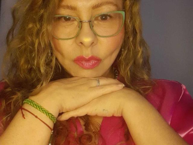 MiaAmoretti with curly brown hair, green glasses, pink lipstick, and pink shirt, pouting with hands under her chin, wearing green and red braided bracelets, and a ring.