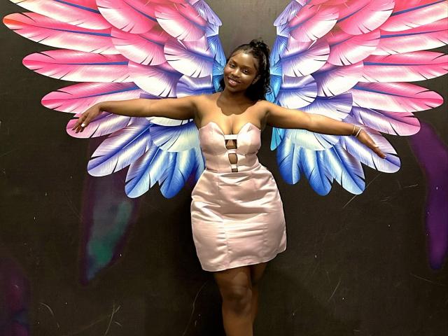Zonniex has big pink and blue feather wings, dark skin, and a cute smile. She's wearing a pink, strapless, cleavage-cut dress with her arms spread wide.