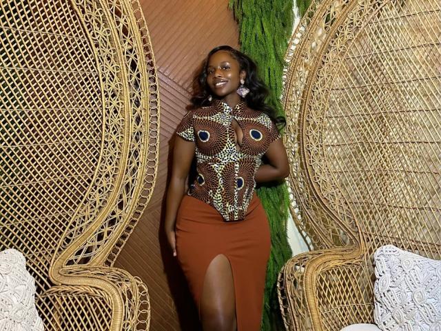 Zonniex has dark skin, big ass, and is wearing a brown, peacock-patterned top with a high slit, brown skirt, and earrings, standing between two wicker chairs.