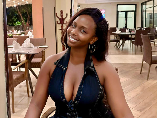 Zonniex has dark skin, medium-sized boobs, and is wearing a black, deep-cut, sleeveless top with zipper, sitting at a table, smiling, with hoop earrings.