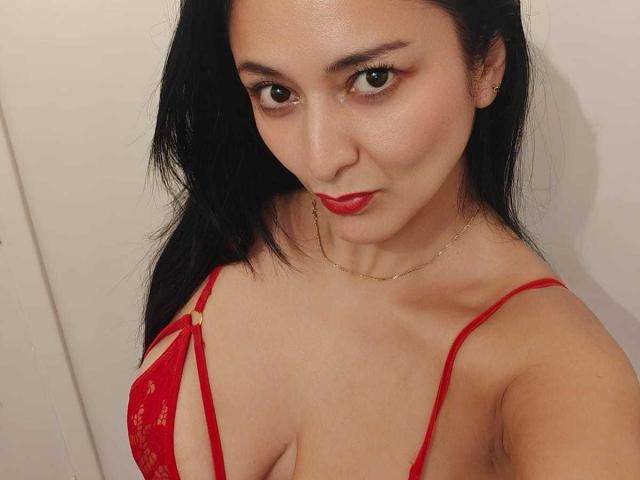 CharlotteGossart with red lace bra, showing her tits, wearing red lipstick, black hair, gold necklace, and a subtle smile, against a plain background.