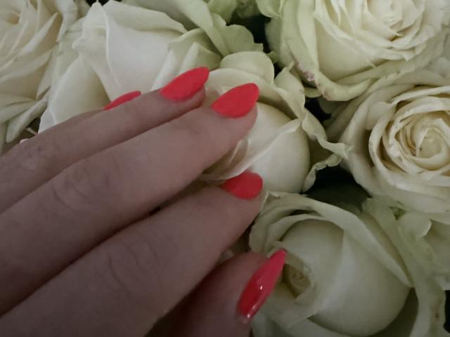 Elionoria's hand with bright red, shiny nails resting on a cluster of white roses. Her fingers are slightly curled, showcasing her long, pointed nails against the soft petals.
