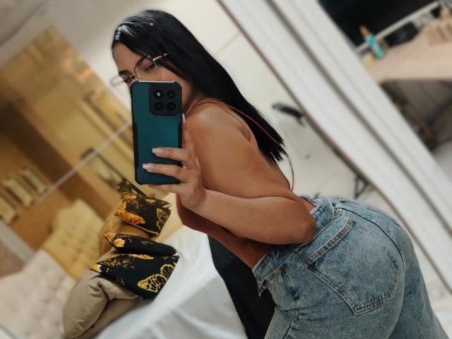 TifaniGreys takes a mirror selfie with her teal phone. She has long black hair, wears glasses, and is topless, exposing her tan skin and small to medium-sized breasts. She's bent over, highlighting her large, round buttocks in blue, high-waisted jeans. The background shows a blurred, beige room with a pillow and a stack of black and yellow pillows on a bed. Her right hand holds the phone, and her left hand rests on her hip. The image has a candid, casual style.