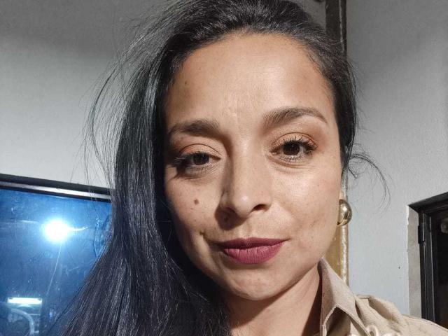 StangelarRivera has dark brown eyes, black hair, and a small mole on her right cheek. She's wearing a beige shirt with pink lipstick and a subtle smile.