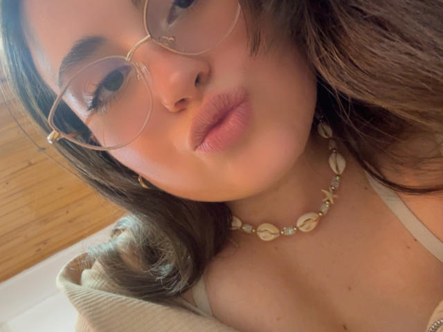 VicluX has puffy pink lips, wearing gold-rimmed glasses, a shell necklace, and a beige top. Her brown hair is wavy, and she's looking at the camera.