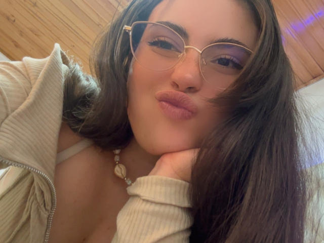 VicluX with pouty lips, wearing gold-rimmed glasses and a beige ribbed jacket, has brown hair, and a white bra is visible, resting her head on her hand.