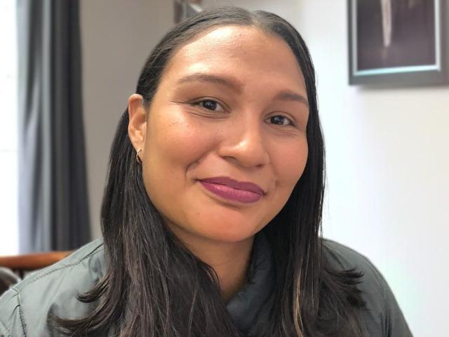 Close-up photo of CoralArzua, a woman with straight, dark brown hair, medium skin tone, and pink lipstick. She wears a green jacket and has small earrings. Her eyebrows are thin, and she has a slight smile. The background is blurred, featuring a dark curtain and a framed picture on a white wall. Her hair is shiny and well-kept, contrasting with her matte skin. The image is well-lit with soft shadows.
