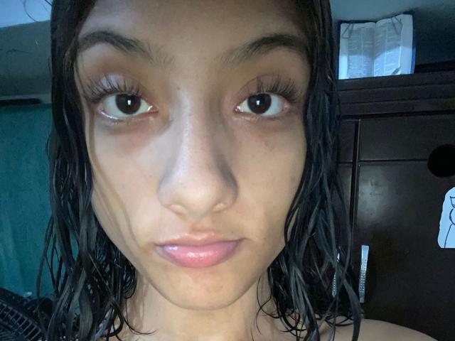 SheaCum's face with wet black hair, brown eyes, and pink lips. Her tits are small, and her pussy is not visible. She has a slight tan and looks serious.