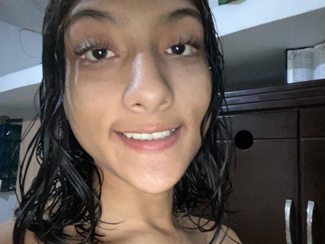 SheaCum's face with wet black hair, brown eyes, small nose, and smiling with white teeth. She has a tear on her right cheek and is looking at the camera.