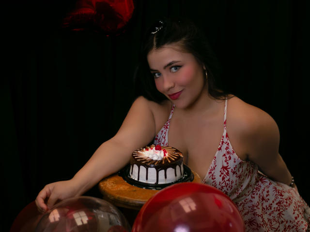 LorensDouce with dark hair, wearing a red floral dress, leaning on a table with a chocolate cake, red balloons, and black background, smiling at the camera.