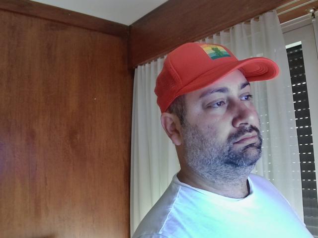 NicoSexHot with a red baseball cap, white shirt, and a slightly messy beard, looking to the side with a neutral expression. His nose is slightly red.