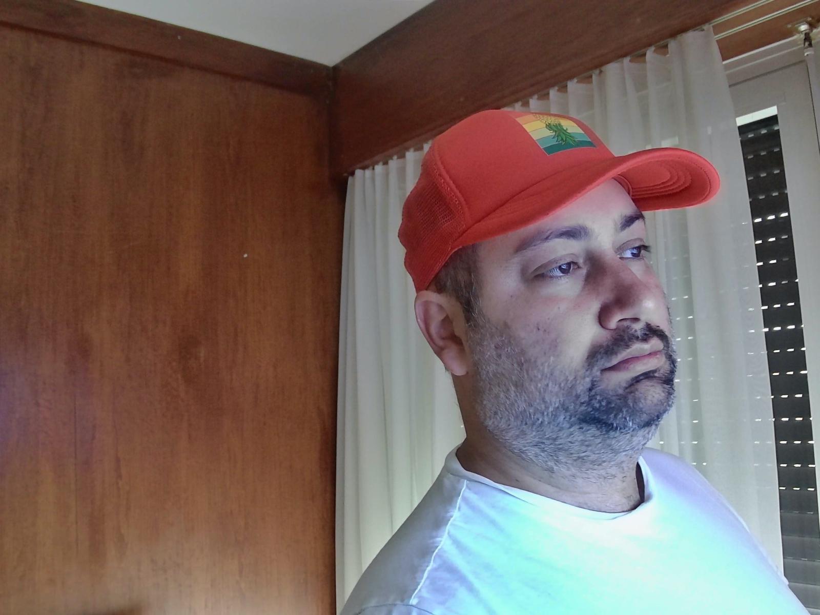 NicoSexHot with a red baseball cap, white shirt, and a slightly messy beard, looking to the side with a neutral expression. His nose is slightly red.
