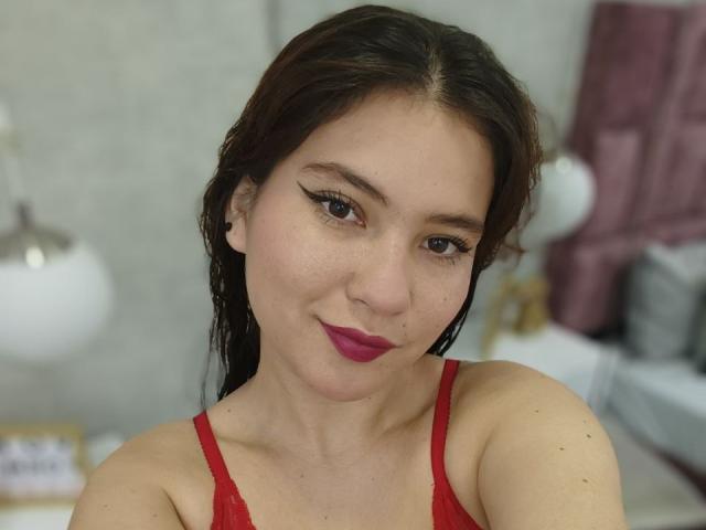 CelesteDash has dark brown hair, red lipstick, and is wearing a red lace bra. She has winged eyeliner, a nose piercing, and is smiling slightly. Her tits are visible.