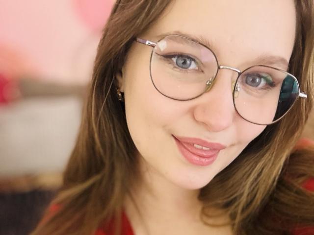 MagikPeachh with glasses, brown hair, and pink lipstick, smiling at the camera. She has blue eyes and a cute, slightly shiny face with subtle makeup.