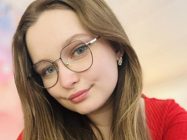 MagikPeachh with long brown hair, wearing glasses and red shirt, has pink lips and small earrings, blue eyes, and a slight smile, looking directly at the camera.