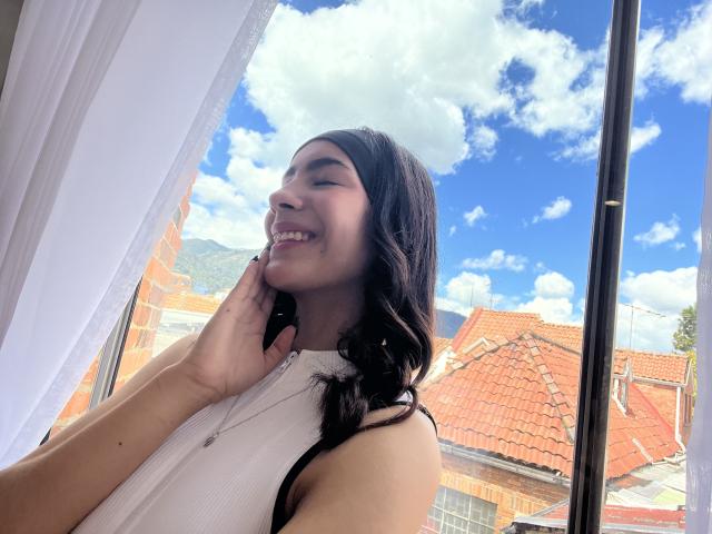 AitanaJohns, smiling, with black hair, wearing a white sleeveless top, touching her cheek, standing by a window, blue sky, white clouds, and red-tiled roofs visible outside.
