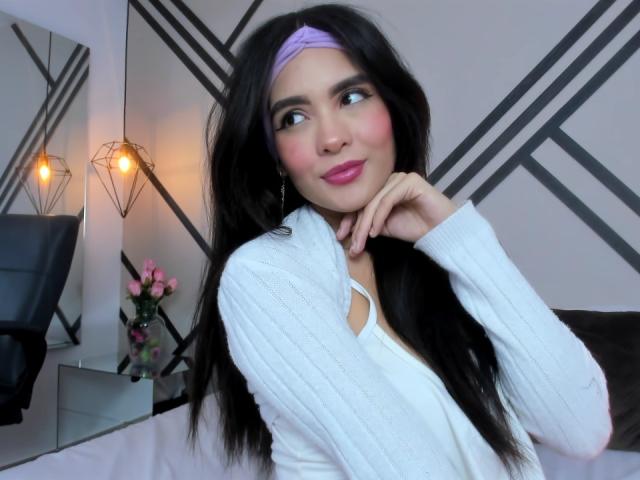 MadisonSmitt with long black hair, pink headband, white rib-knit top, pink lipstick, and blushing cheeks, resting her chin on her hand, smiling gently.