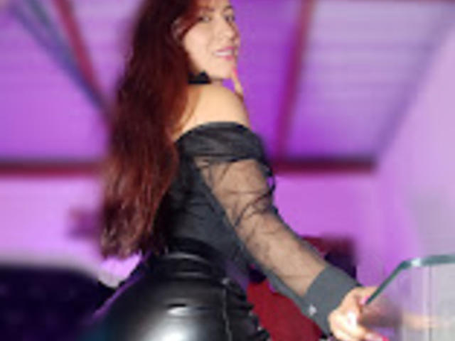 SofiaFountaine with long red hair, black off-shoulder top, and tight black leather pants, smiling, holding a green microphone, looking over her shoulder.