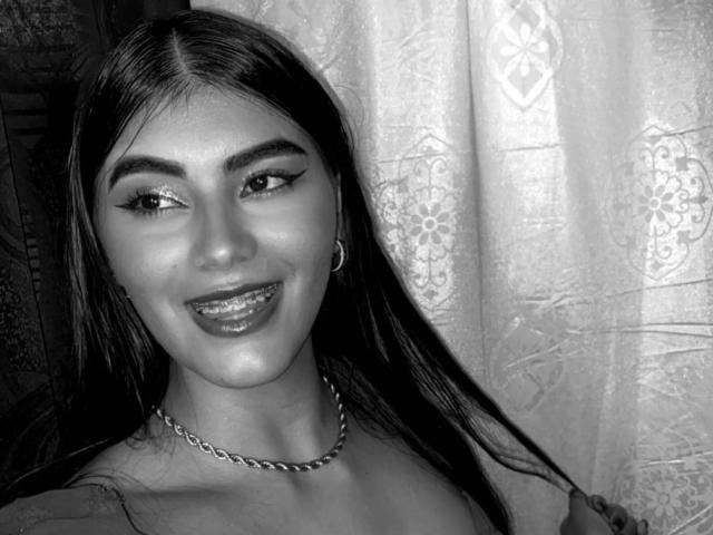 Abby Kell tem olhos brilhantes e cintilantes, um sorriso adorável com lábios brilhantes, e cabelos longos e escuros. Ela está usando um colar de corrente e brincos, e parece sexy em preto e branco..