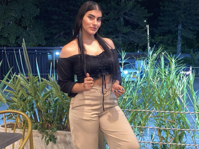 Abby Kell, met lang donker haar, een zwarte top met een open schouder en beige broek met hoge taille, staat, geeft een duimpje omhoog, toont haar borsten, in een buitenomgeving 's nachts, met planten..