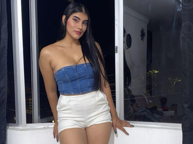 AbbyKell with long black hair, wearing a blue strapless denim top and white high-waisted shorts, standing in a window at night, showing her tits and ass.