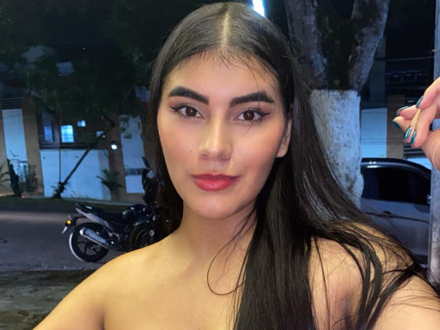 Abby Kell, avec ses longs cheveux noirs, ses yeux marrons et son rouge à lèvres rouge, dévoilant ses épaules nues, et un léger sourire, avec une main levée, dans un décor de rue nocturne..