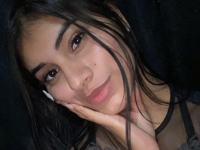 Abby Kell tem olhos castanho-escuros, cabelo preto e uma pequena marca de beleza na bochecha direita. Ela está tocando o rosto com a mão direita, vestindo uma blusa preta transparente..