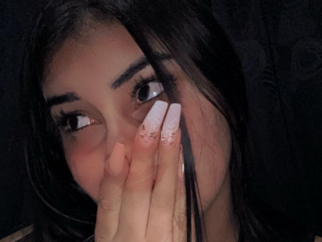 Os olhos castanho-escuros de Abby Kell olham para o lado, cobrindo a boca com uma mão que exibe unhas longas, pintadas de branco com pontas rosa. Cabelo preto, levemente bagunçado, em primeiro plano..