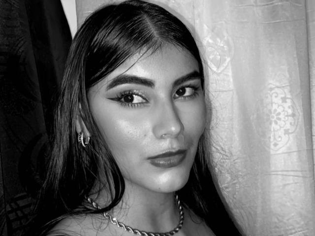 Abby Kell heeft donker, steil haar, opvallende eyeliner en een lichte glimlach. Ze draagt een ketting en kleine oorringen. Haar huid is glad en licht glanzend..