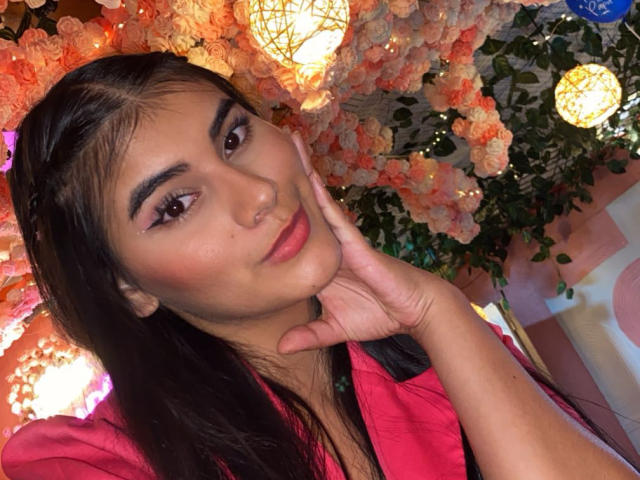 Abby Kell, avec de longs cheveux bruns tressés sur le côté, un chemisier rose, le menton appuyé sur sa main, des lèvres roses, des yeux marrons, un maquillage discret, un sourire doux, entourée de guirlandes lumineuses chaudes et de fleurs roses..