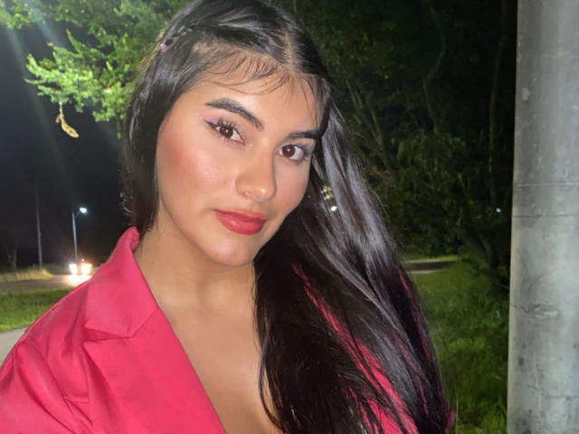 Abby Kell, com seus longos cabelos pretos, batom vermelho e sombra rosa, vestindo uma camisa vermelha, tem um rosto adorável e olhos castanhos sensuais. Seus seios estão levemente visíveis..