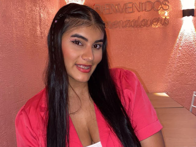 Abby Kell, met lang zwart haar, rode lippenstift en een roze shirt, glimlacht naar de camera en laat haar borsten zien in een top met een lage halslijn. Ze heeft donkere ogen en een schattig gezicht..