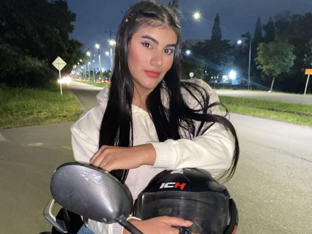 AbbyKell stands in a nighttime urban setting, holding a black motorcycle helmet with "IC" in red letters. She has long black hair, dark eyebrows, and wears a white hoodie. Her right hand is on the motorcycle's handle, and her left arm rests on the helmet. The image is lit with streetlights, creating a contrast with her dark hair and white hoodie. The road and grassy area are visible in the background.
